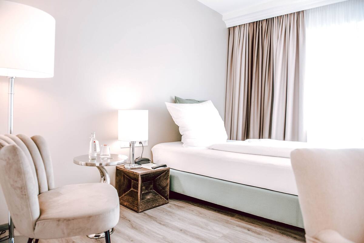 A hotel room with a bed, a bedside table with a lamp, a chair, and a small wooden side table.