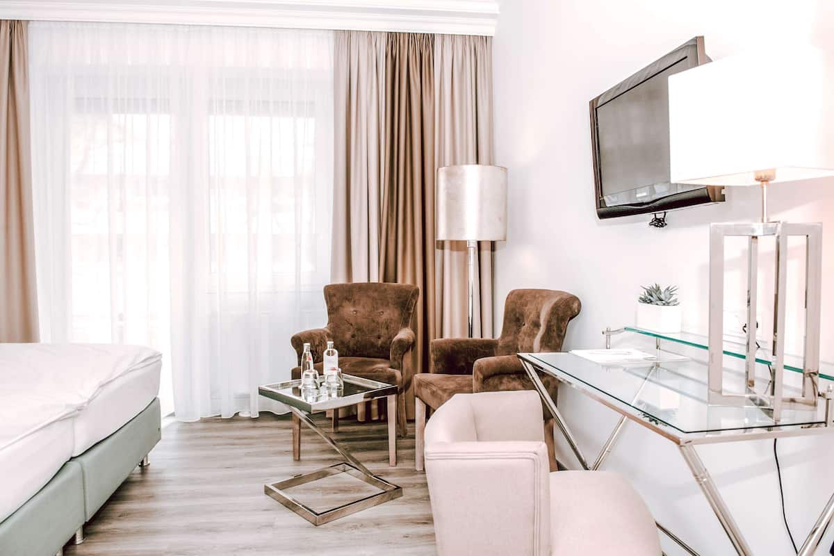 A hotel room with a bed, two armchairs, a glass coffee table, a desk with a lamp, a TV mounted on the wall, and a window with curtains.