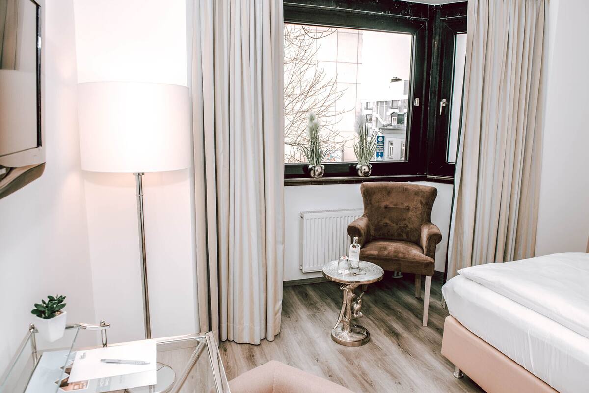 A hotel room with a bed, a chair, a lamp, a small table with a bottle, and a window with curtains.