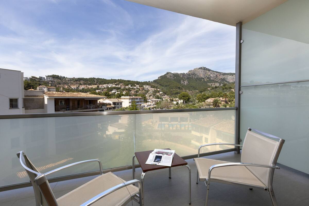 A balcony with two chairs, a small table with a newspaper, and a view of a town and mountains.