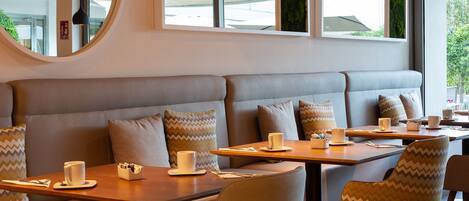 A modern cafe with a seating area featuring cushioned benches and tables, a round mirror, and large windows with a view of the outdoors.