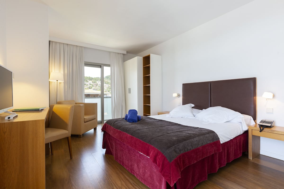 A modern hotel room with a large bed, a desk, a chair, a TV, and a window with curtains.