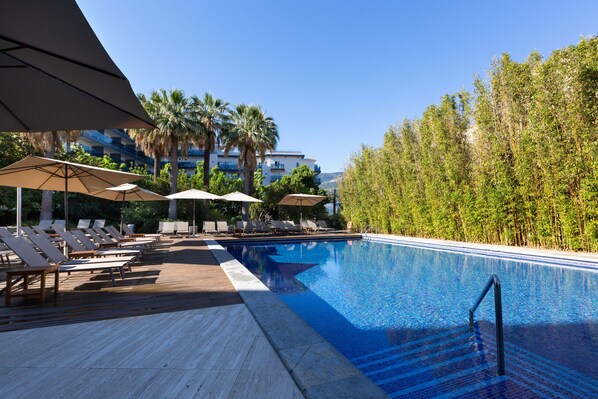 Uma área de piscina com espreguiçadeiras e guarda-sóis, cercada por vegetação e um edifício ao fundo.