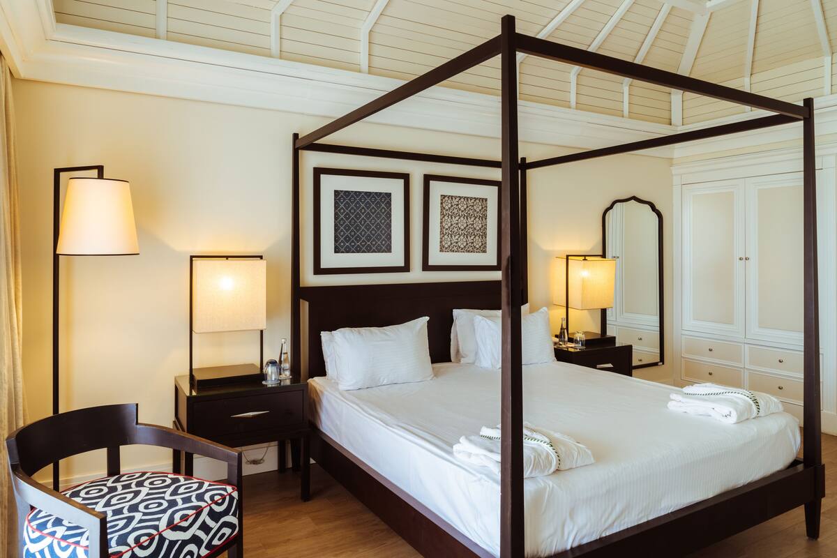 A four-poster bed with white linens, a wooden nightstand with a lamp, a chair with a patterned cushion, and framed artwork on the wall.