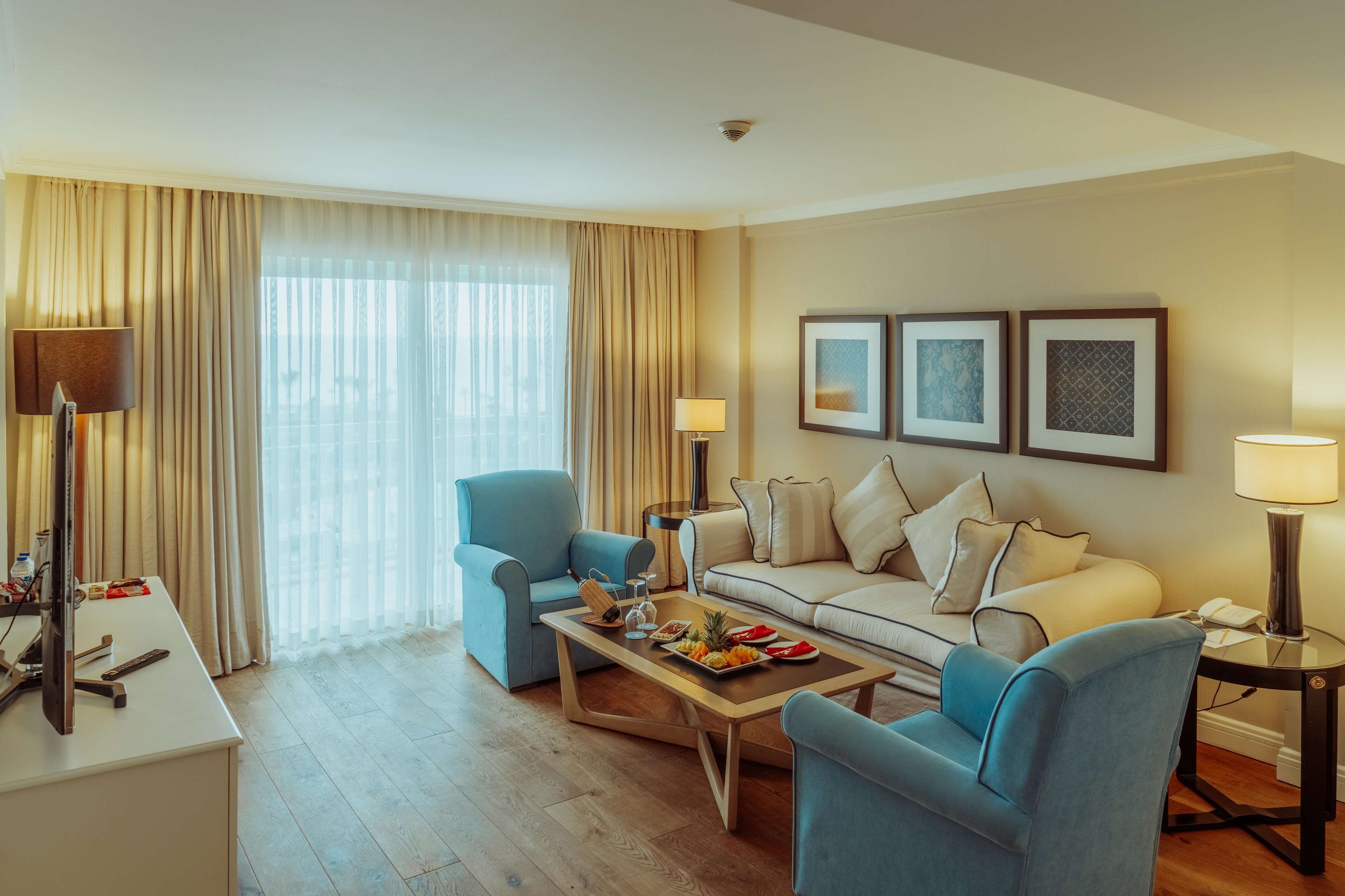 A modern hotel room with a sofa, two armchairs, a coffee table with fruit, a desk with a computer, and a large window with curtains.