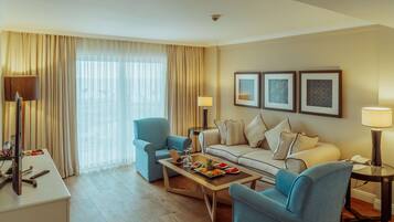 A modern hotel room with a sofa, two armchairs, a coffee table with fruit, a desk with a computer, and a large window with curtains.