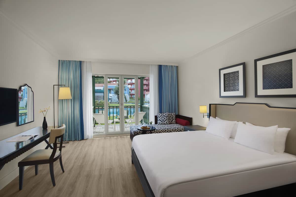 A hotel room with a large bed, a desk with a chair, a TV, and a balcony with a pool view.