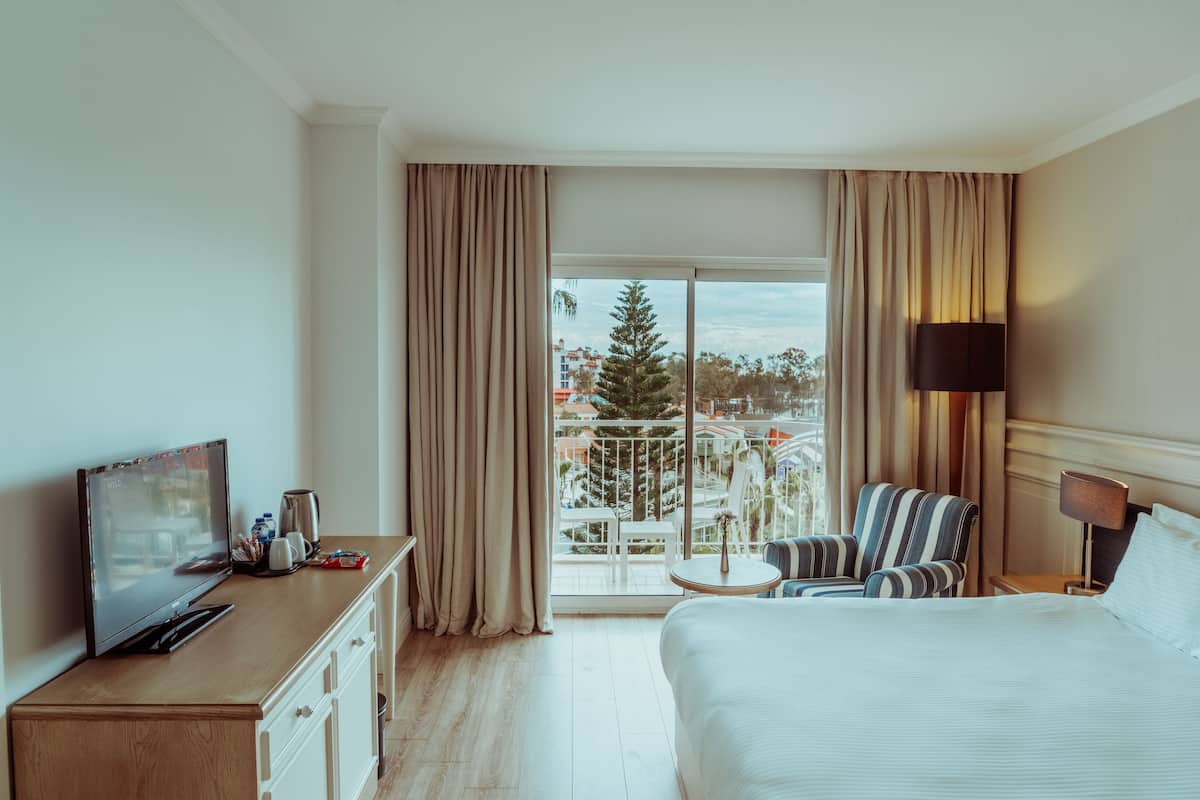 A hotel room with a bed, a TV, a desk with a coffee maker, a chair, and a balcony with a view of buildings and trees.
