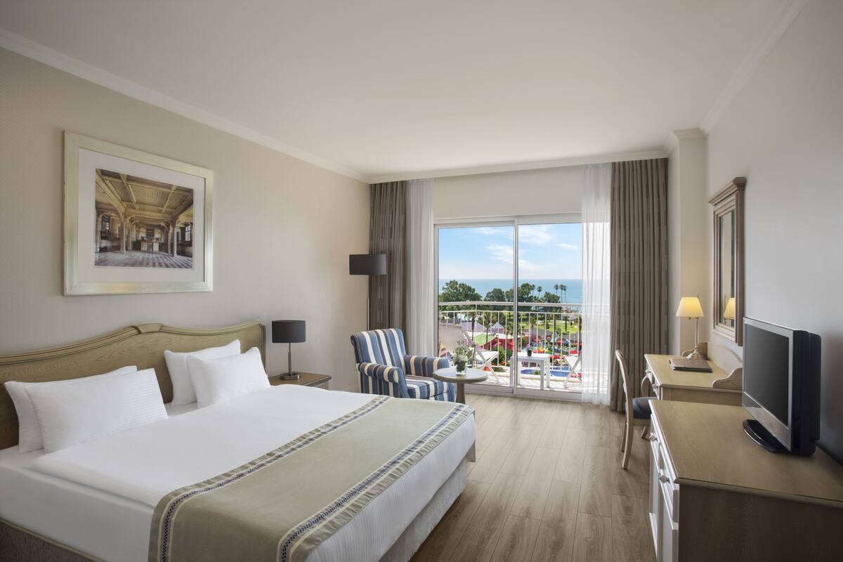 A hotel room with a large bed, a desk, a chair, a TV, and a balcony with a view.