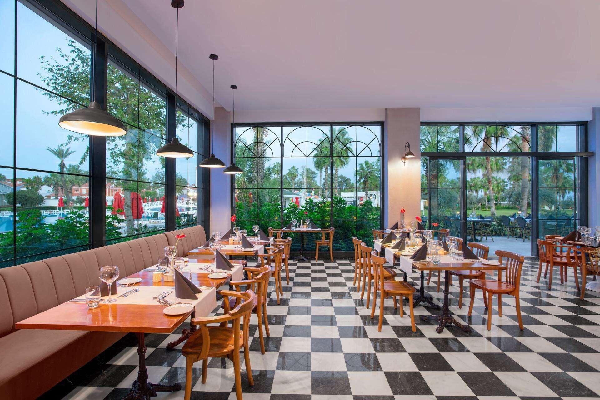 Un restaurant au sol à carreaux, avec des tables et des chaises en bois. De grandes fenêtres offrent une vue sur des palmiers et une piscine.