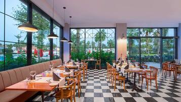 Un restaurant au sol à carreaux, avec des tables et des chaises en bois. De grandes fenêtres offrent une vue sur des palmiers et une piscine.