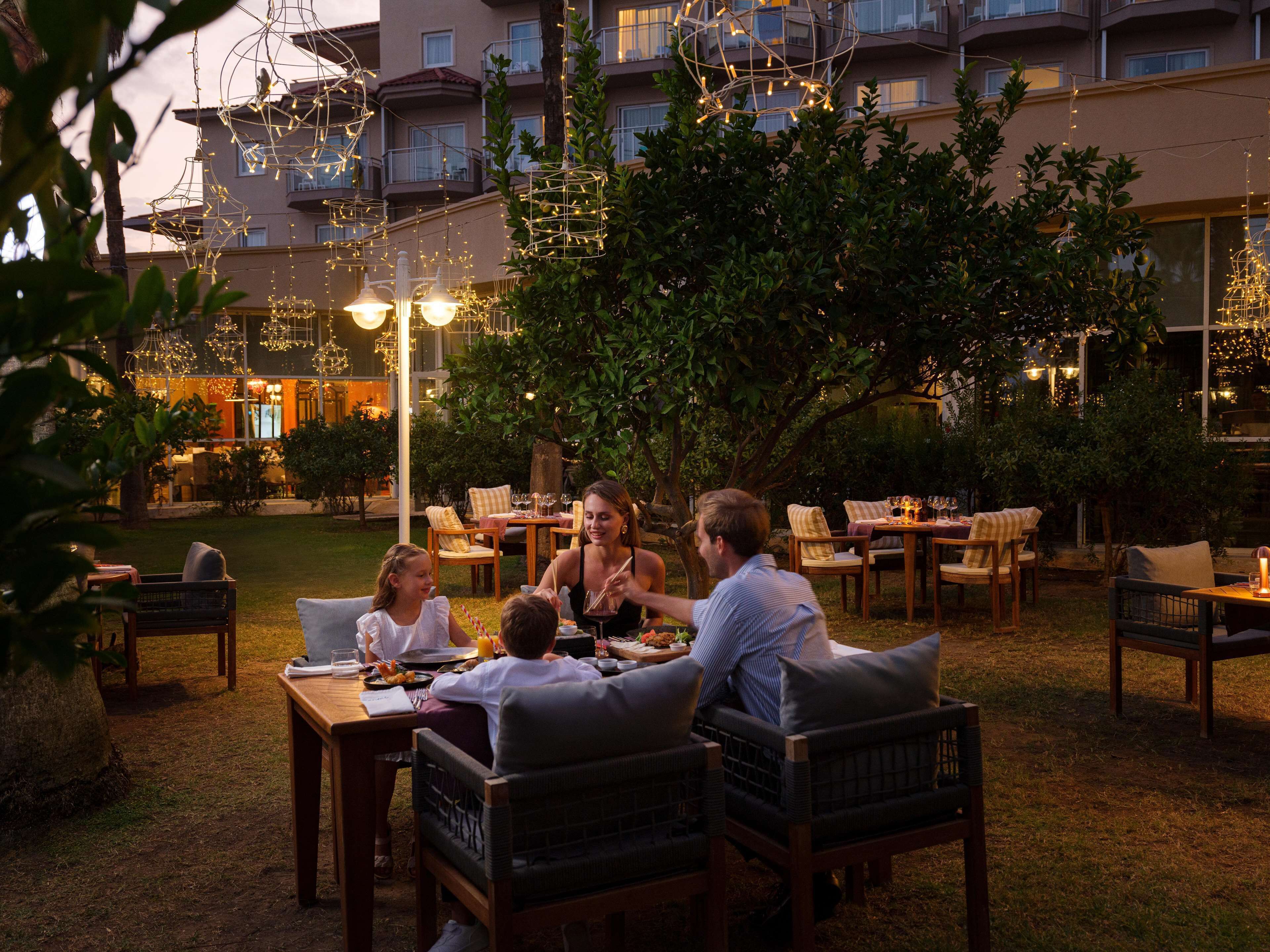 An outdoor dining area with tables and chairs, surrounded by greenery and illuminated by string lights.