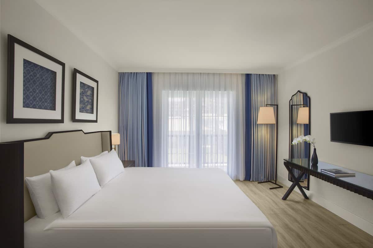 A hotel room with a large bed, a desk with a lamp, a TV mounted on the wall, and two framed pictures on the wall.