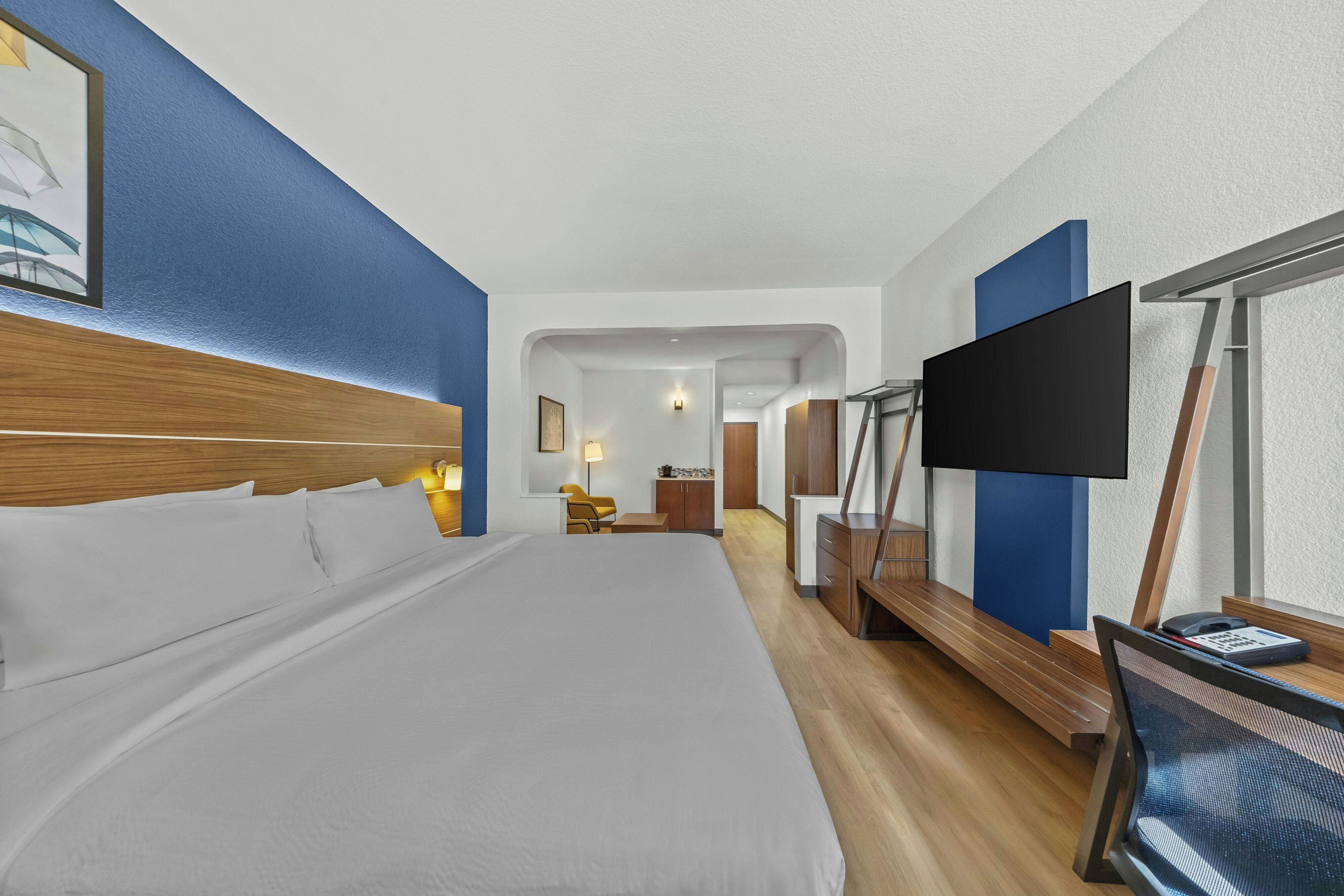 Habitación de hotel con una cama grande, un escritorio con una silla, televisor y baño visible a través de un arco.