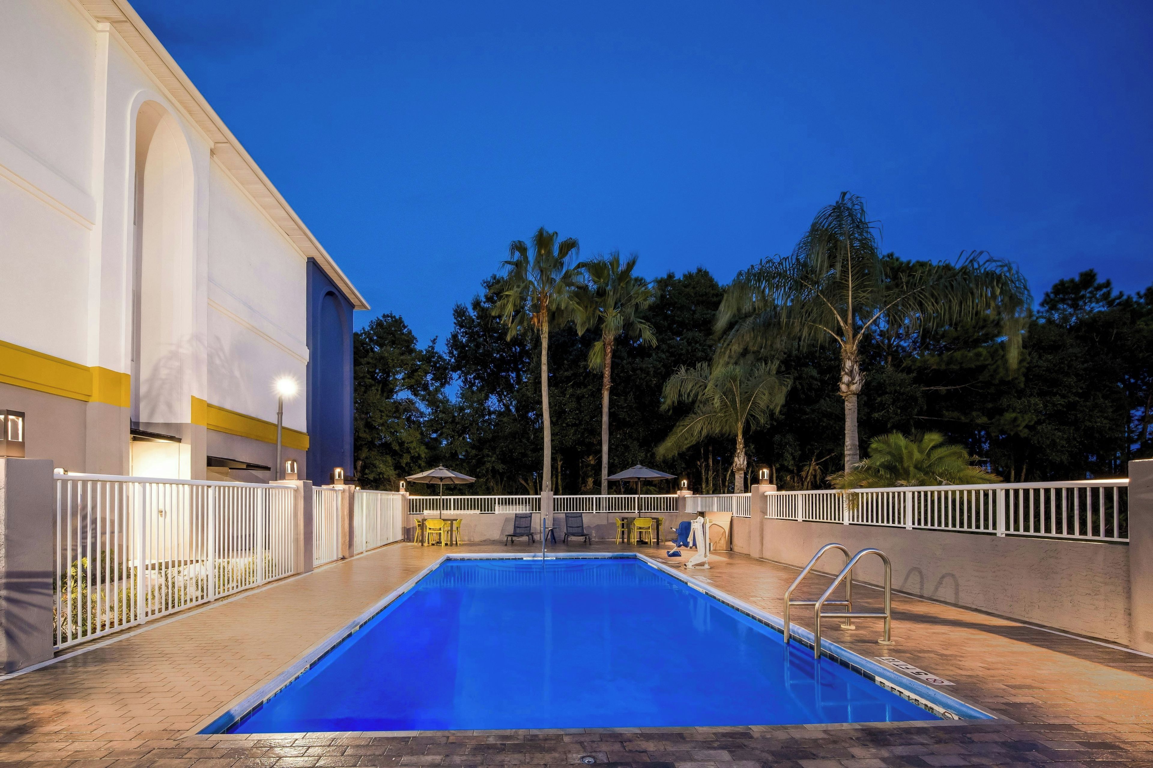 Una piscina con valla blanca, palmeras y un edificio con una franja amarilla.