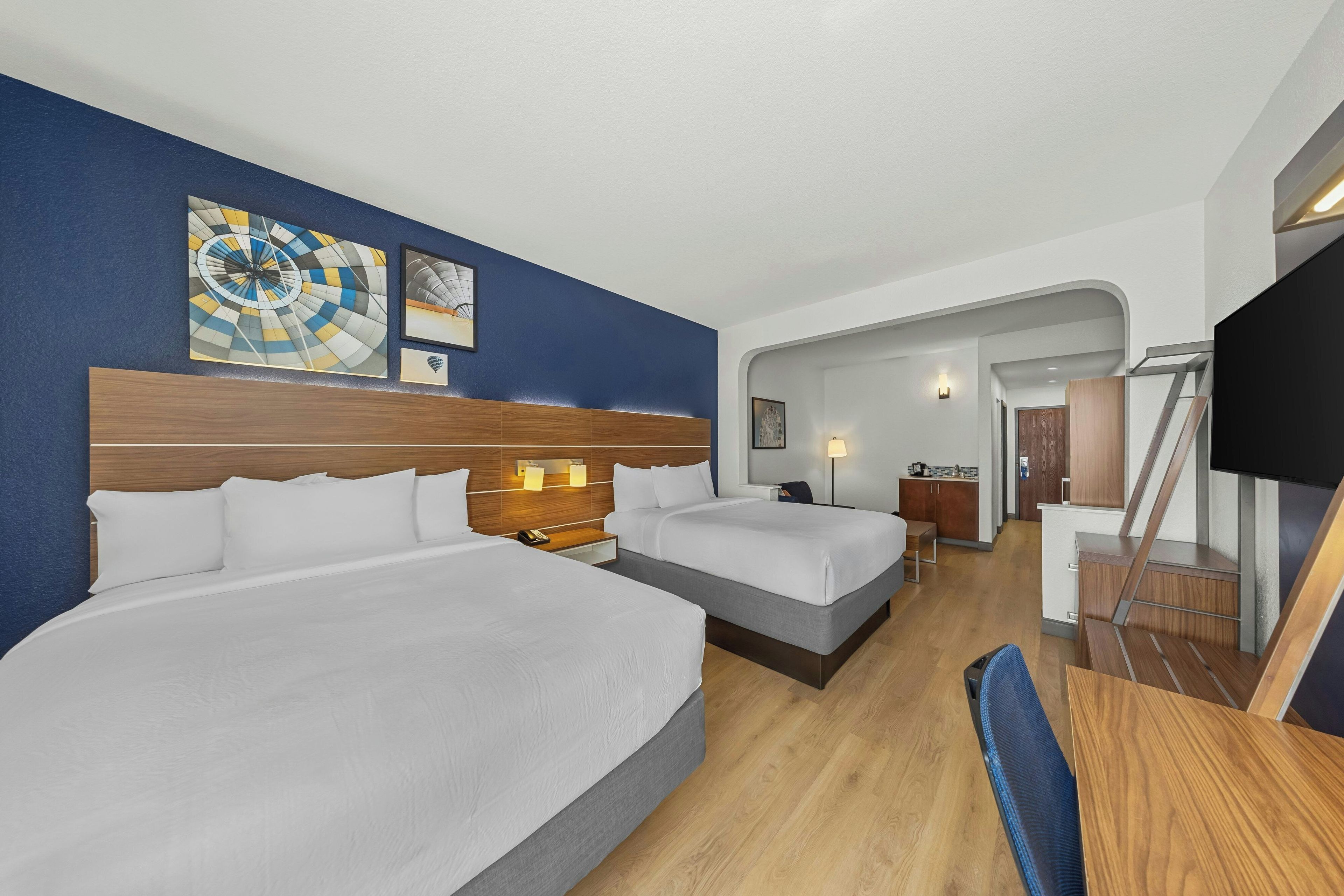 A hotel room with two beds, a desk, a chair, a TV, and artwork on the walls.