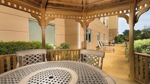Un patio couvert avec une table ronde et des chaises, des balustrades en bois et une vue sur un bâtiment doté de fenêtres et bordé d’une haie.