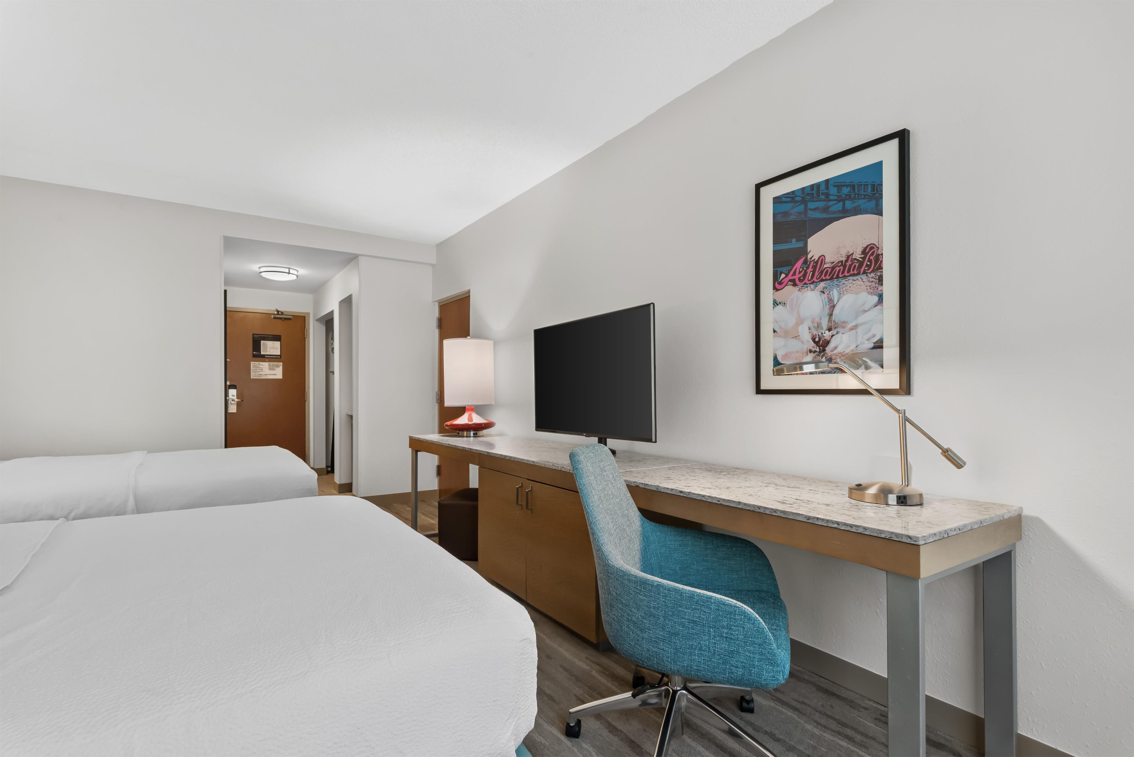 A hotel room with a bed, a desk with a computer, a chair, and a poster on the wall.