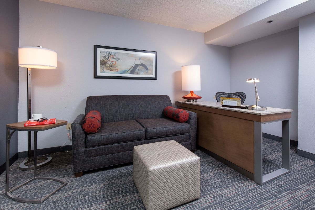 A hotel room with a gray sofa, a small ottoman, a desk with a lamp, and a framed artwork on the wall.