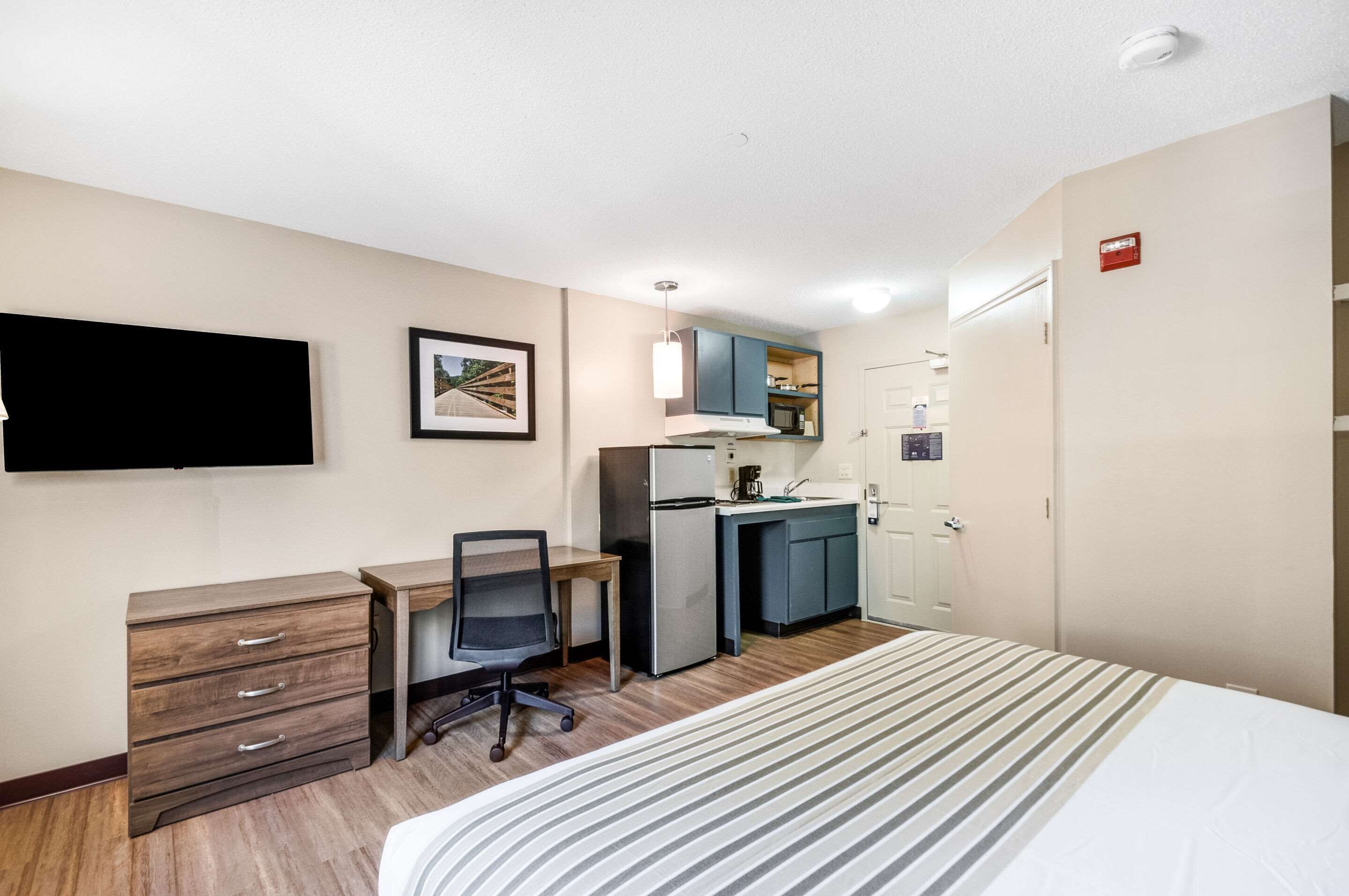 Habitación de hotel con cama, escritorio con silla, kitchenette, televisión y un cuadro en la pared.