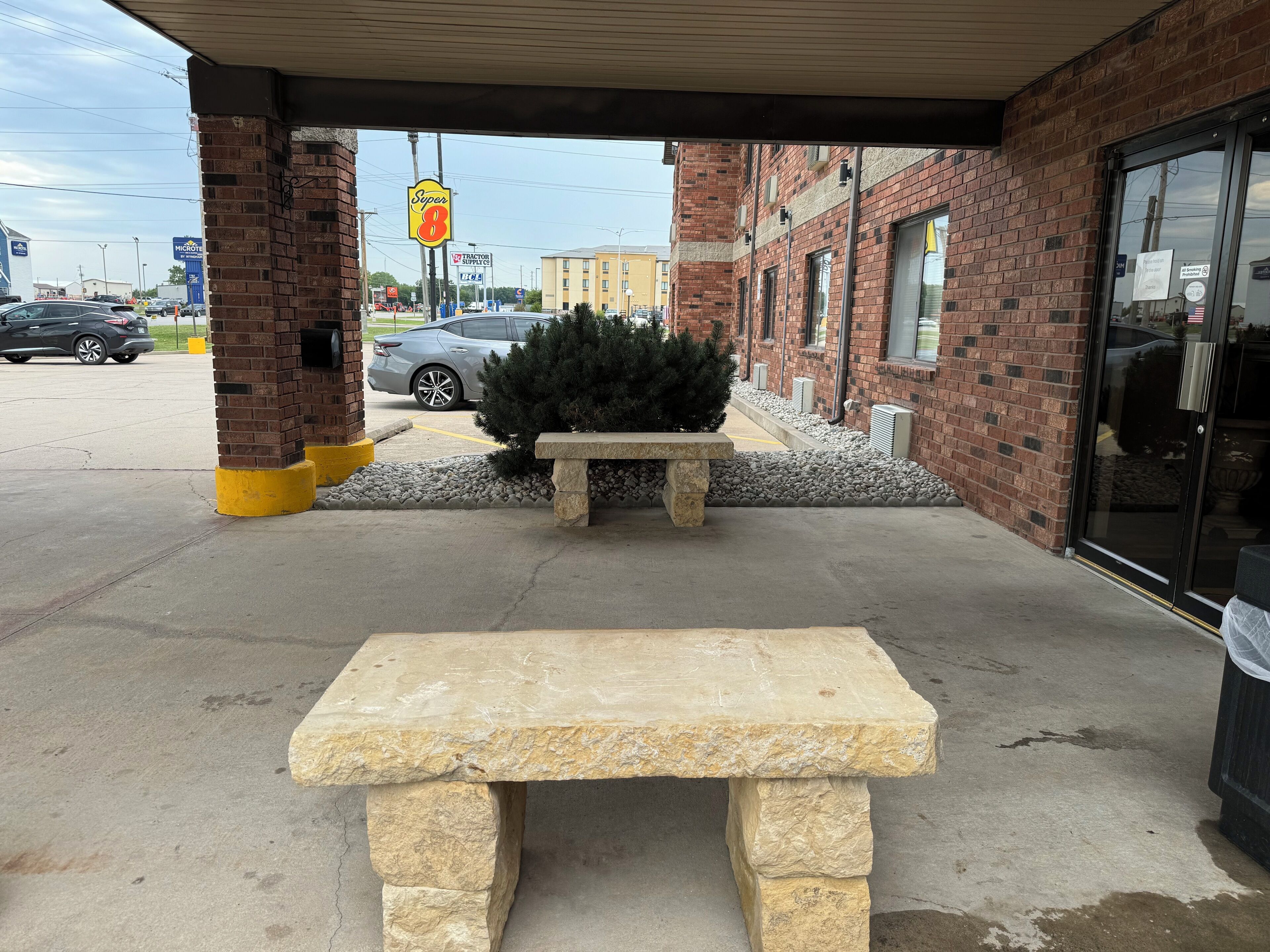 Un banco de piedra bajo una zona techada, con un edificio de ladrillo al fondo.