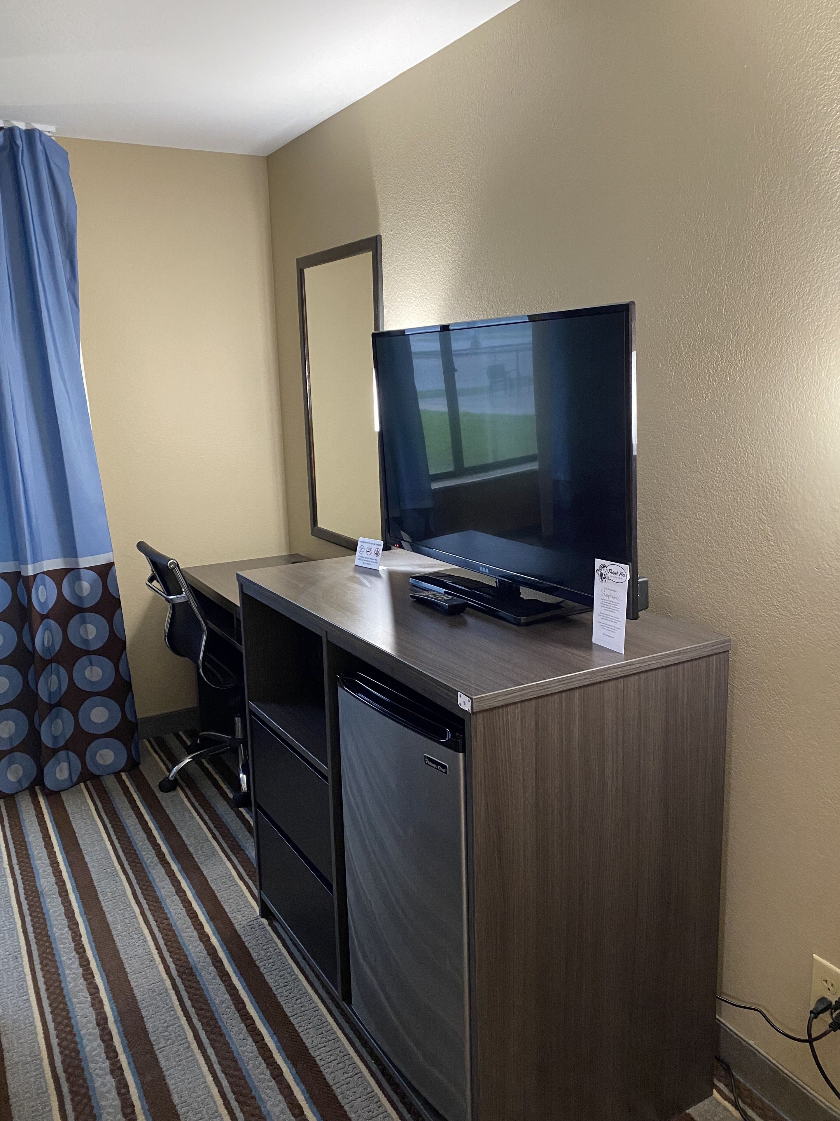 A hotel room with a wooden desk, a black monitor, a keyboard, and a small refrigerator.
