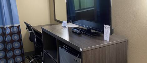 A hotel room with a wooden desk, a black monitor, a keyboard, and a small refrigerator.