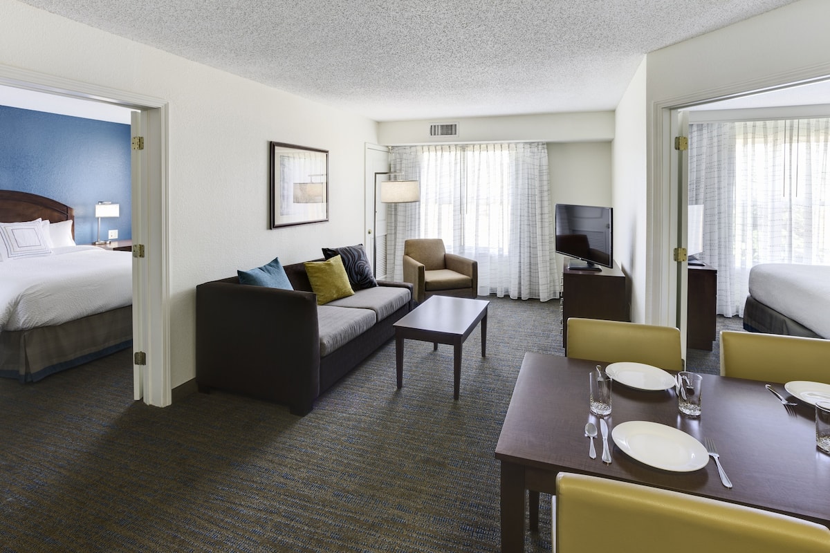 A hotel room with a bed, a sofa, a dining table set with plates and cutlery, a TV, and a lamp.