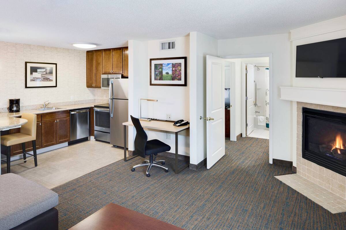 A compact hotel room with a kitchenette, a dining area, a workspace, and a fireplace.