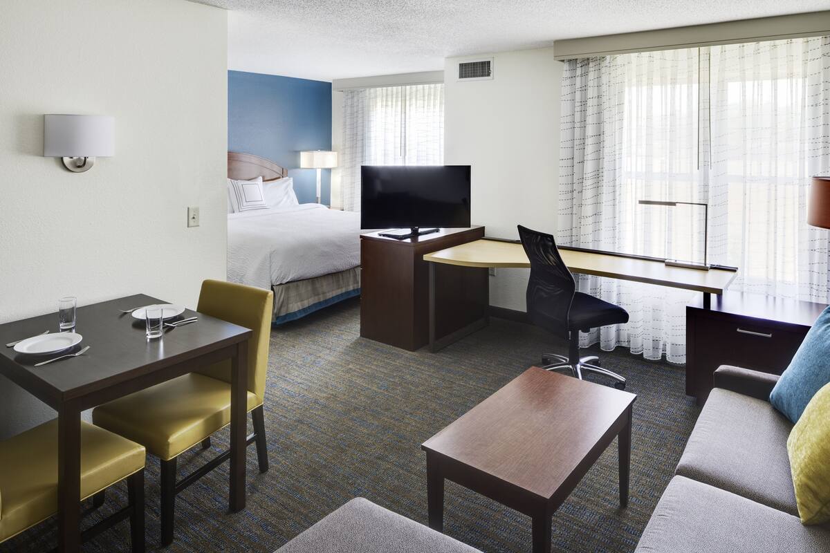 A hotel room with a bed, a desk with a TV, a dining table with chairs, and a sofa.