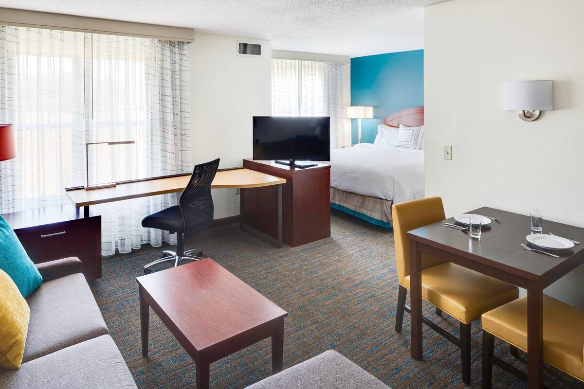 A hotel room with a bed, a desk with a TV, a sofa, a dining table with chairs, and a carpeted floor.
