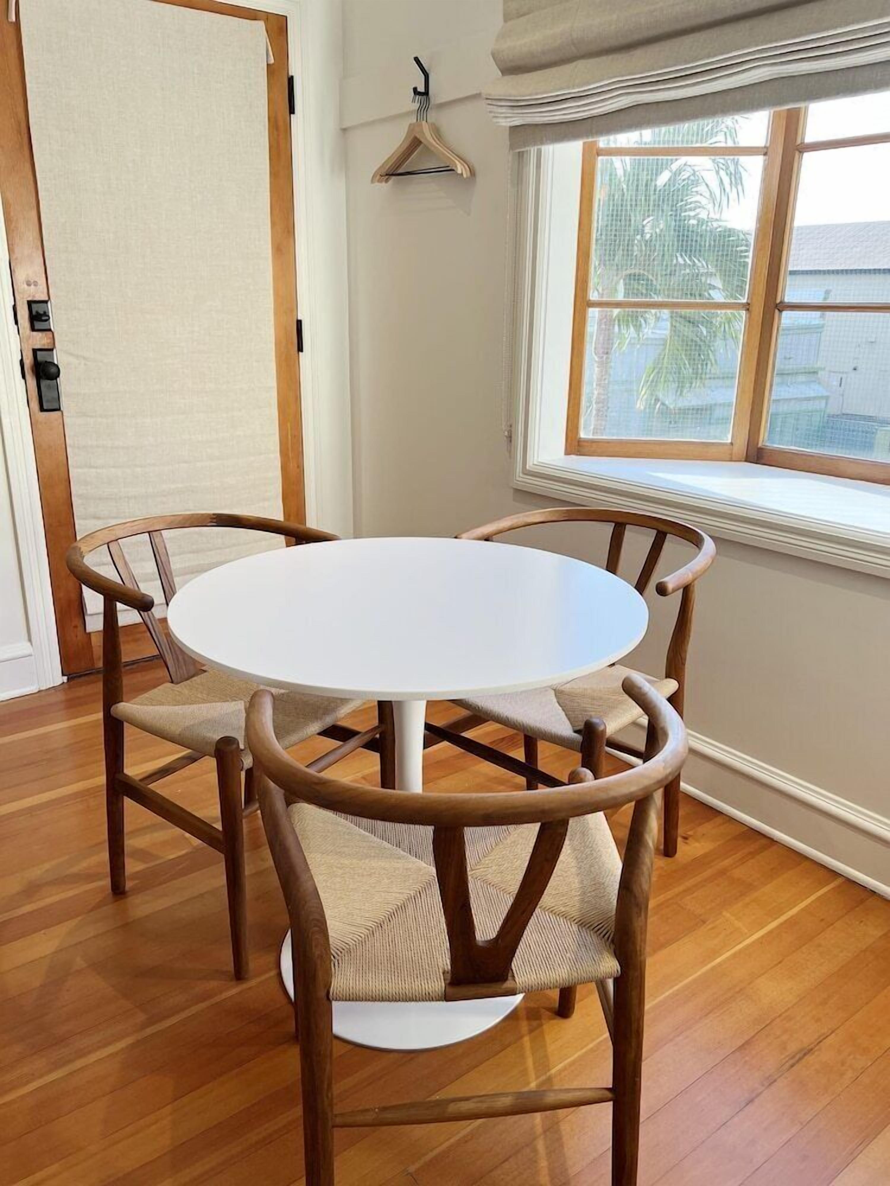A round dining table with four wooden chairs, a wooden door, a window with a view of palm trees, and a wall-mounted hanger.