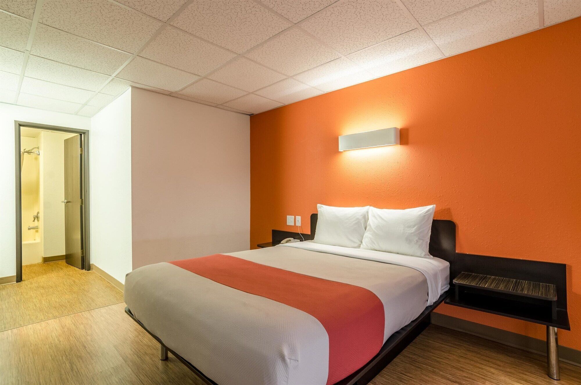 A hotel room with a bed, a nightstand, a wall-mounted light, and a door leading to a bathroom.