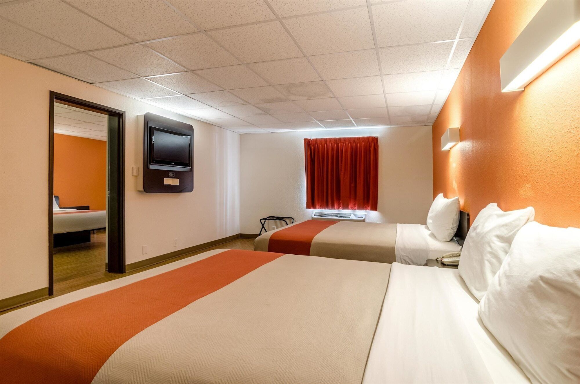 Habitación de hotel con dos camas, un televisor, una ventana con cortinas rojas y una puerta que da a otra habitación.