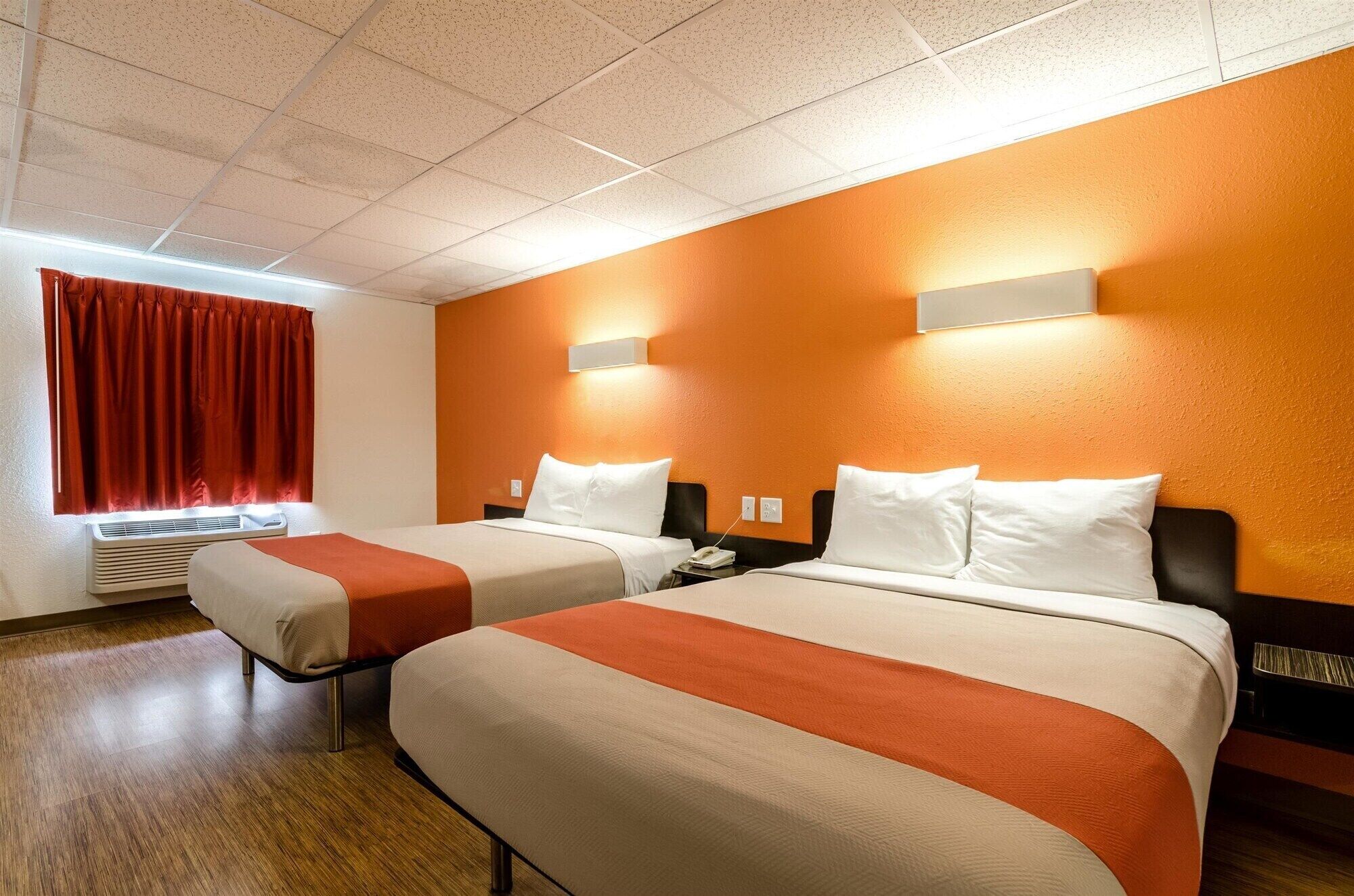 Quarto de hotel com duas camas, janela com cortinas vermelhas, unidade de ar condicionado e luminária fixada na parede.