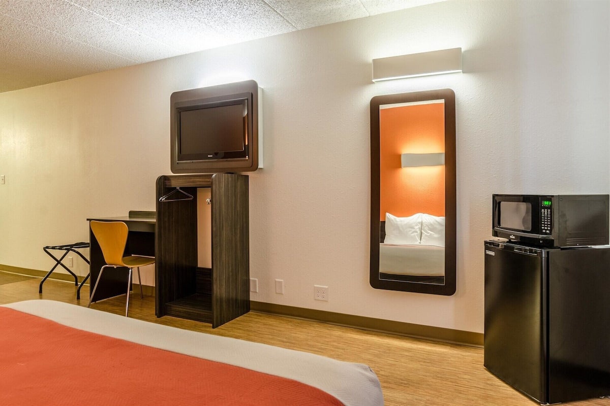 Une chambre d’hôtel comprenant un lit, un téléviseur fixé au mur, un four à micro-ondes, un petit bureau avec une chaise et un miroir.