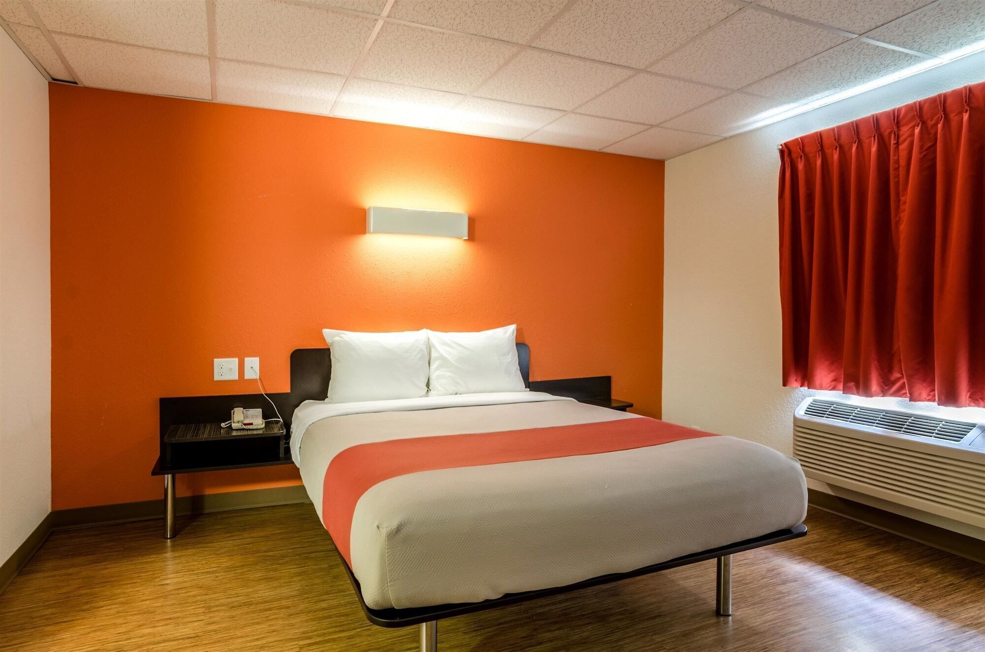 Une chambre d’hôtel avec un mur d’accent orange, un lit avec une literie blanche, une table de chevet avec un téléphone et une fenêtre avec des rideaux rouges.