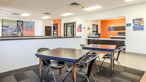 A hotel reception area with a counter, two tables, and chairs. There are signs on the wall and a digital screen. - Motel 6 York, NE (York)