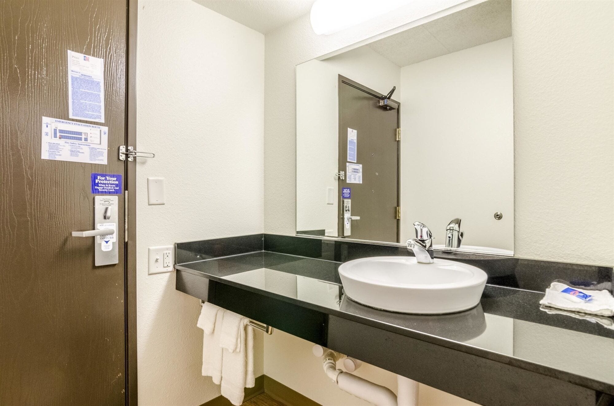 Baño de hotel con encimera negra, lavamanos blanco, espejo grande y toallero.