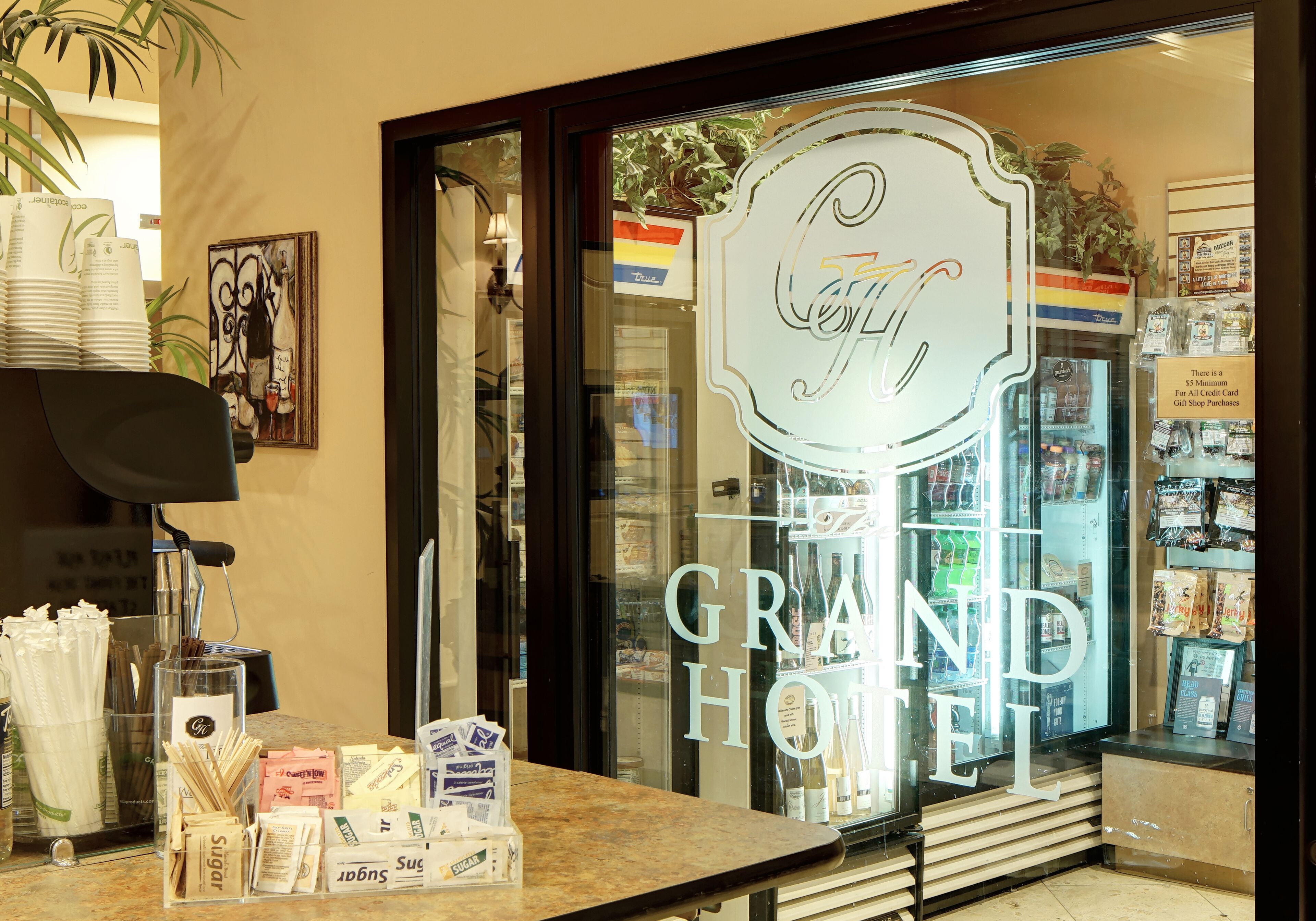 A hotel reception area with a glass front displaying the name 'Grand Hotel'. There are stacks of white cups and saucers, a glass display case with various items, and a sign indicating a minimum spend for credit card purchases.
