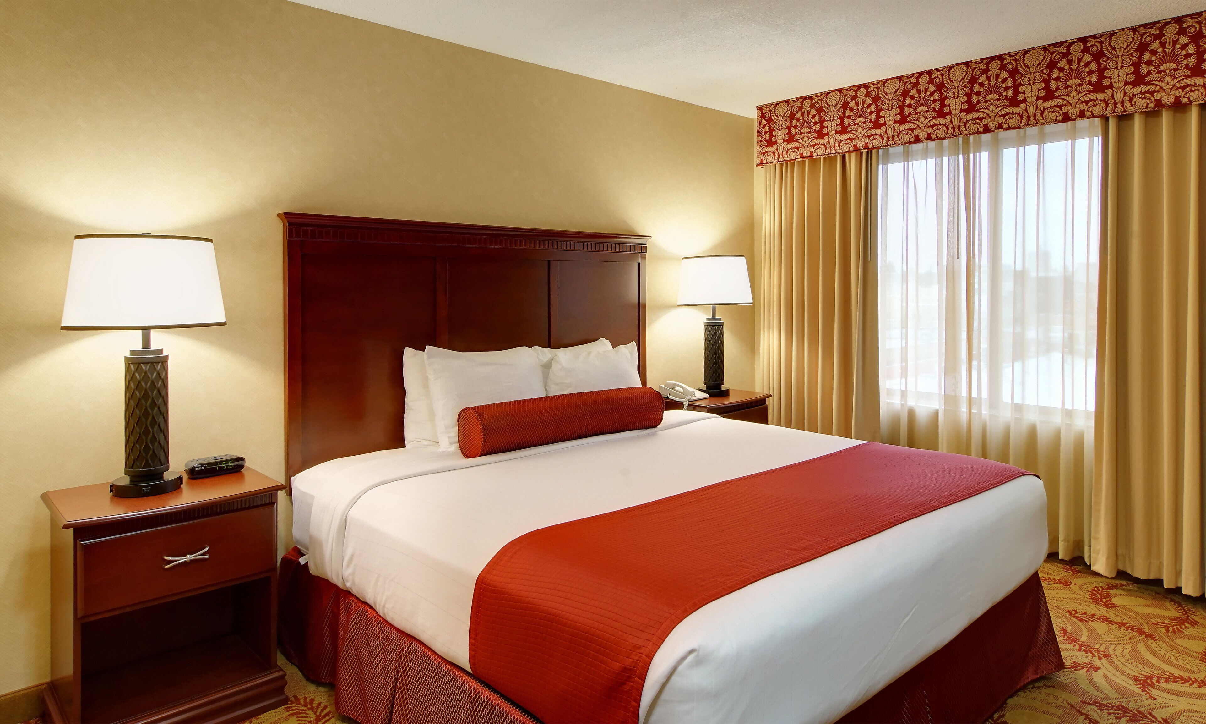A hotel room with a large bed, two bedside tables with lamps, a wooden headboard, and a window with curtains.