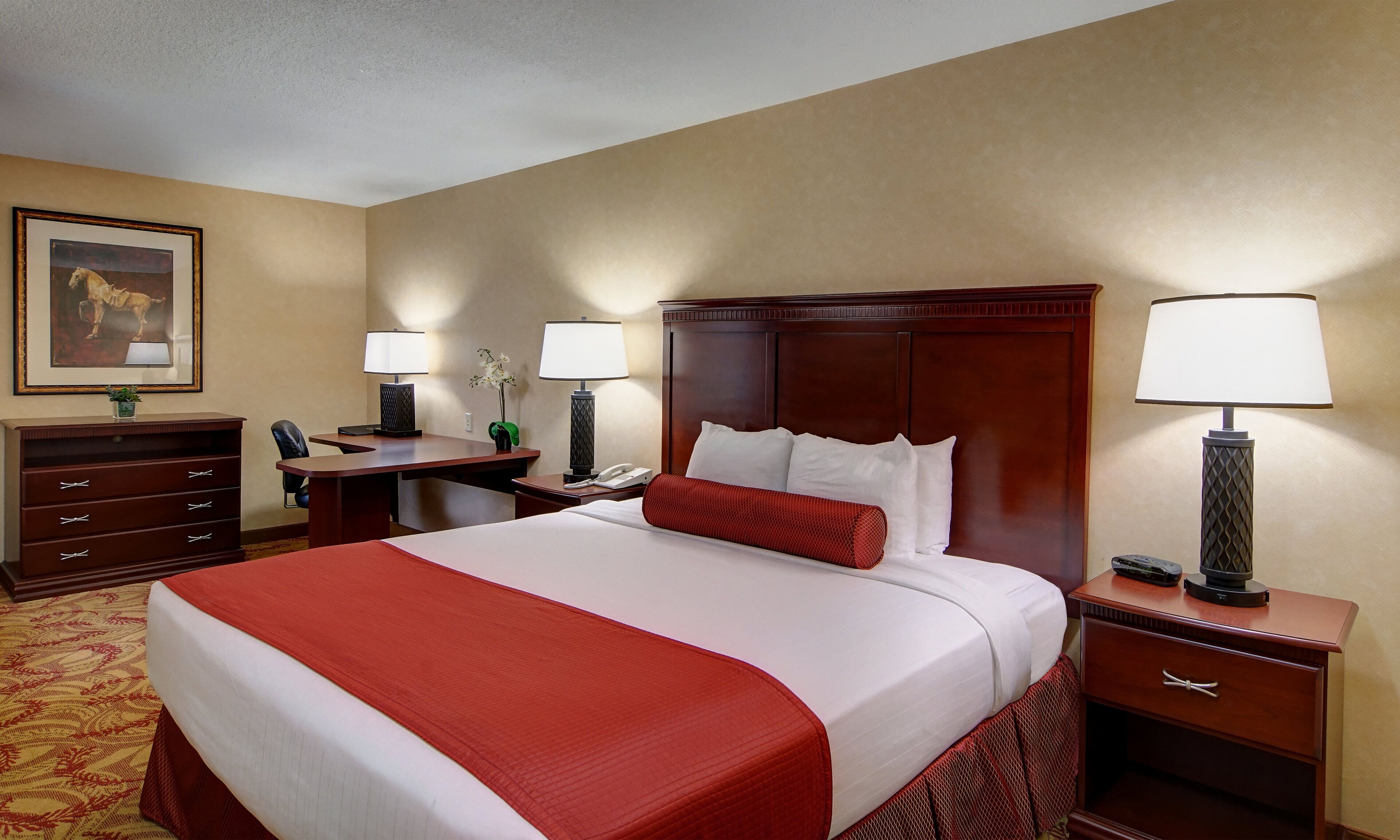 A hotel room with a large bed, a desk with a lamp, a telephone, and a small plant.