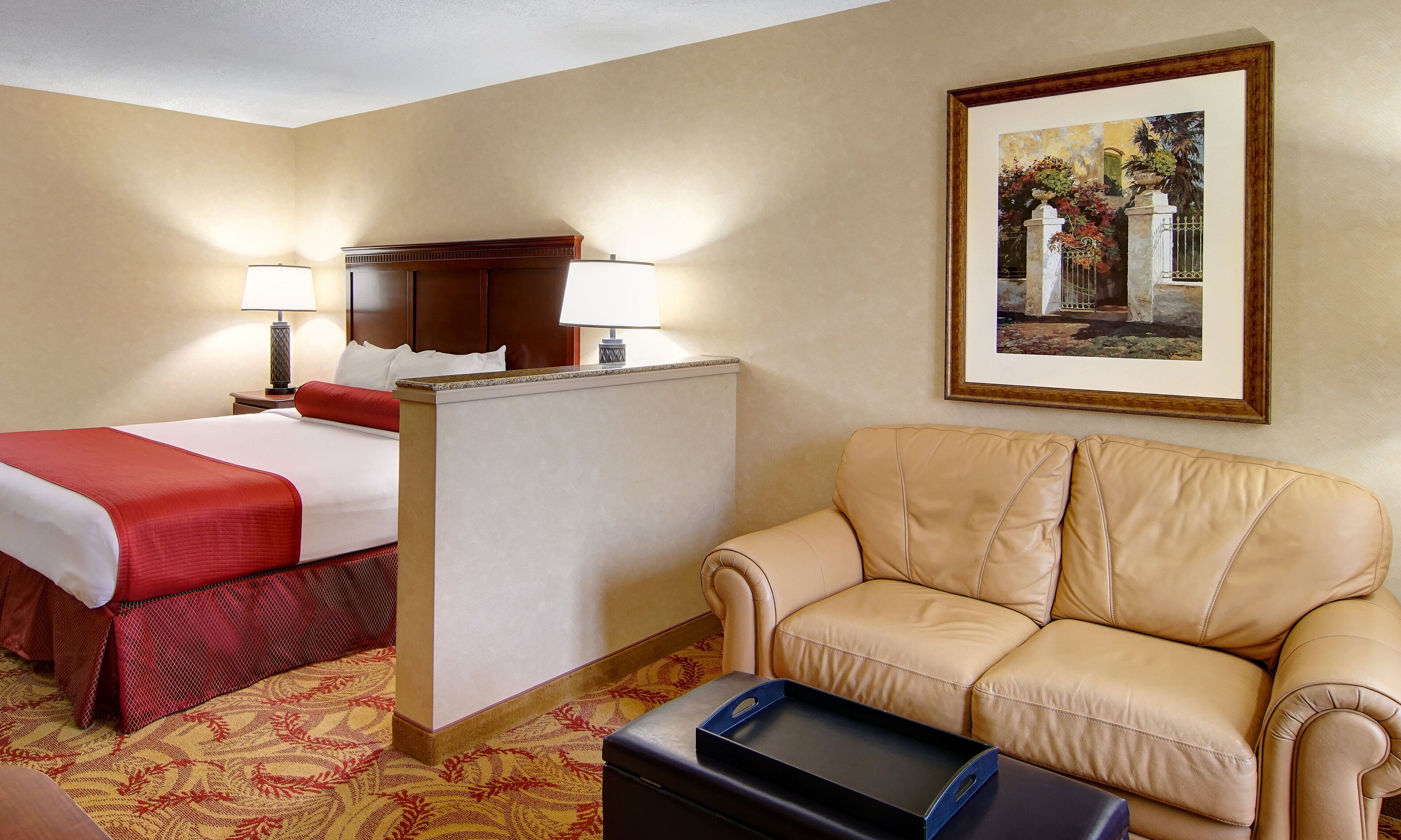 A hotel room with a bed, a sofa, a desk with a lamp, and a framed picture on the wall.