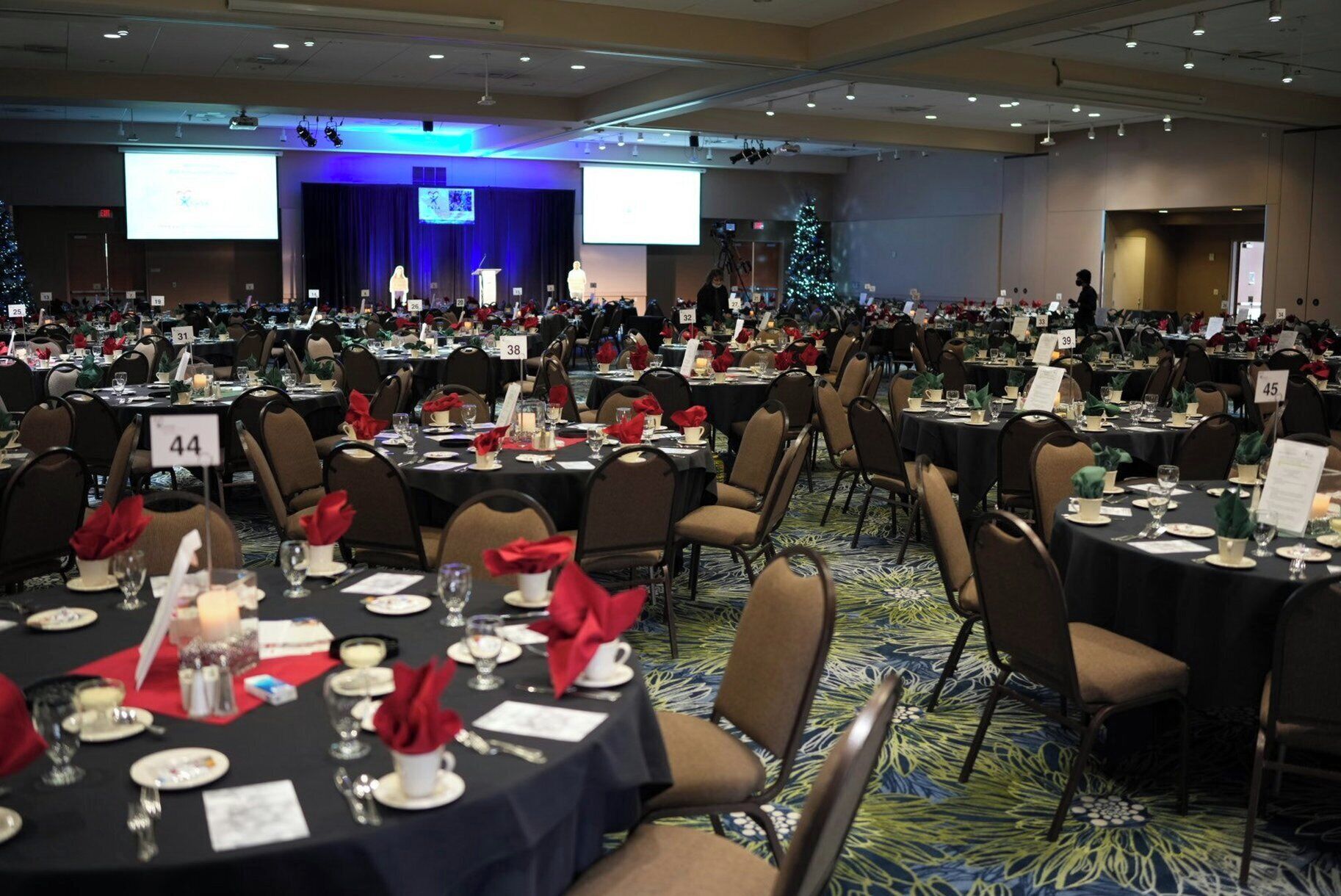 Un salón de banquetes con mesas redondas, sillas numeradas y un escenario con pantalla.