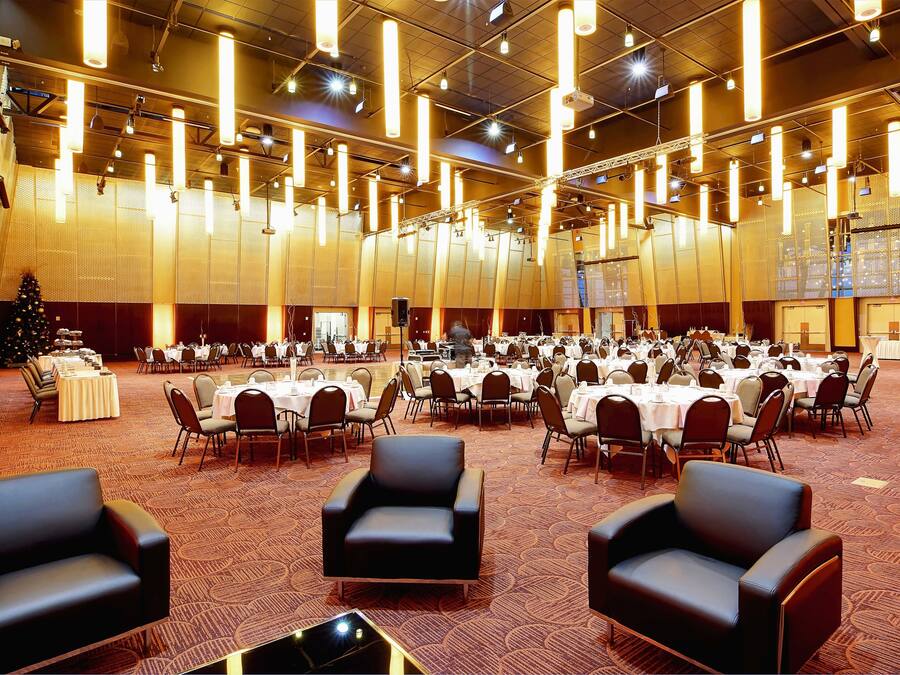 A large banquet hall with round tables and chairs, high ceiling with hanging lights, and a Christmas tree.