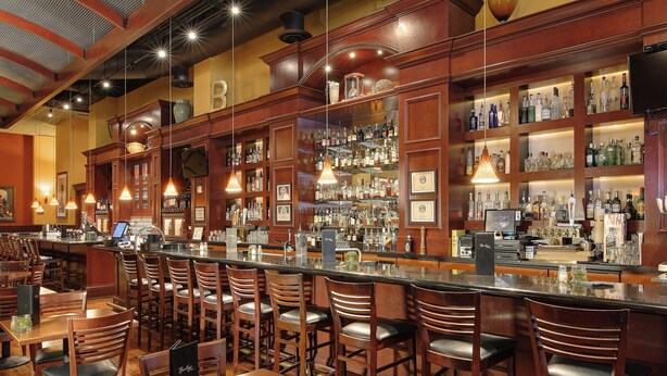A bar area with wooden chairs and tables, a bar counter, and shelves stocked with bottles and glasses.