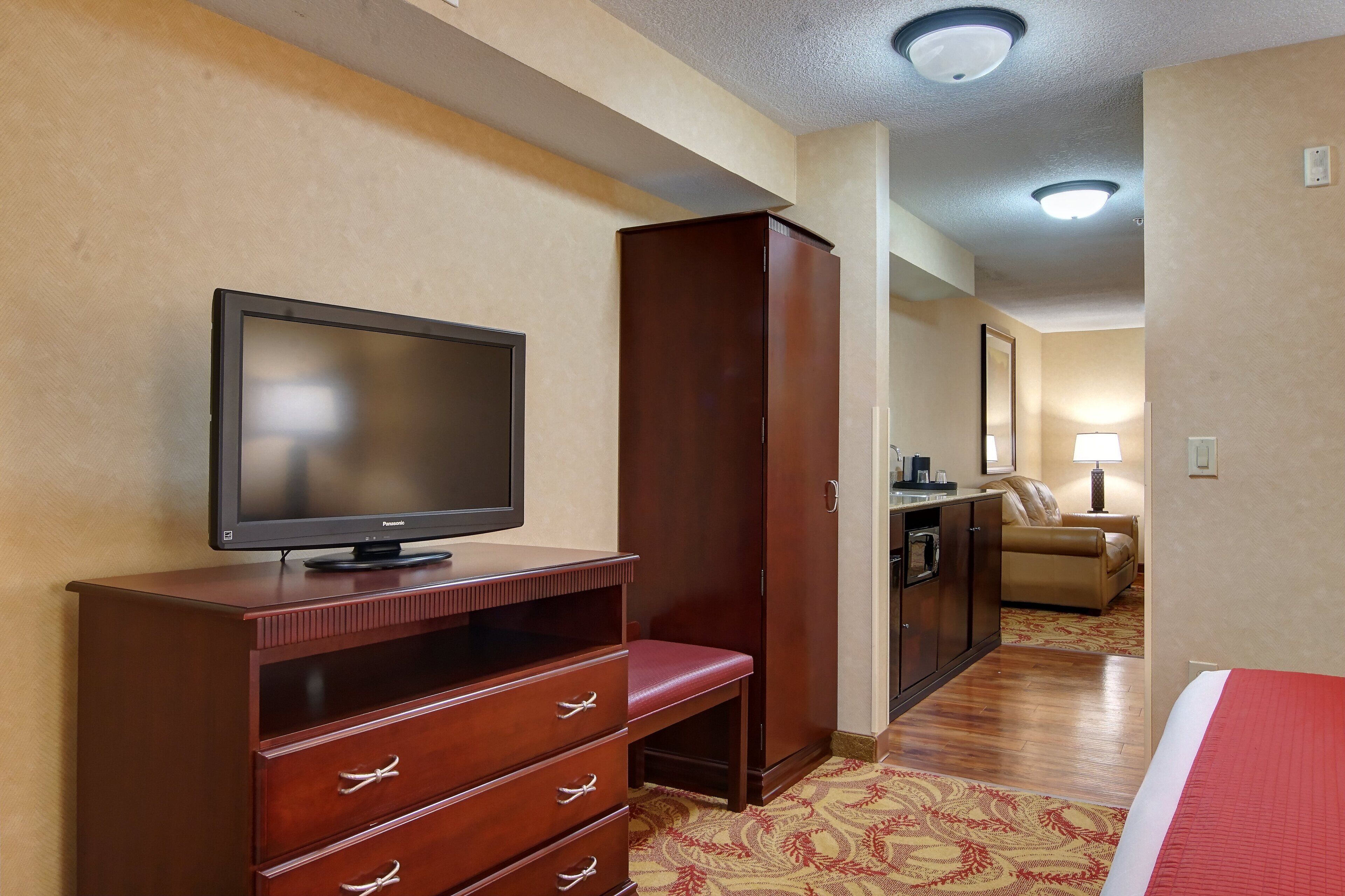 Quarto de hotel com televisão sobre um armário de madeira, guarda-roupa, cama com manta vermelha, sofá e tapete estampado.