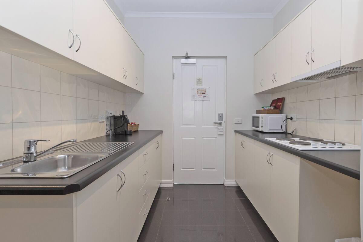 A narrow kitchen with white cabinets, a stainless steel sink, a microwave, and a refrigerator.