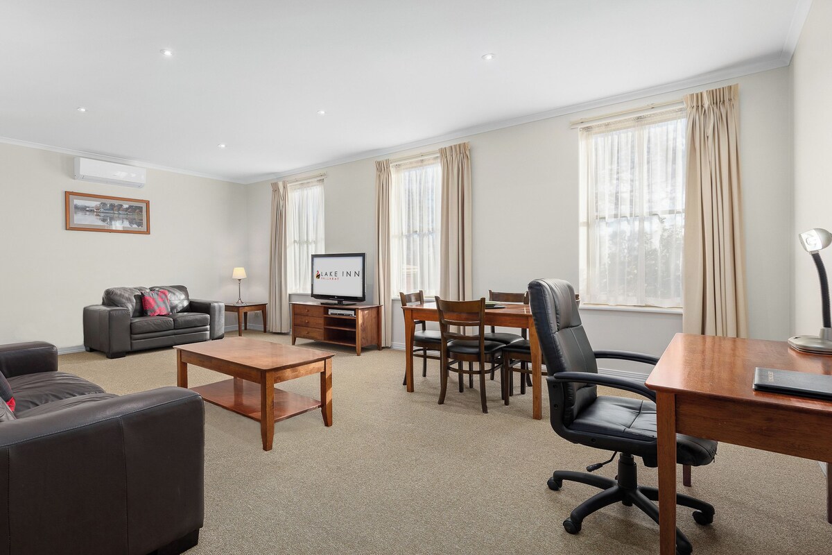 A hotel room with a living area, a dining table, a sofa, a TV, and a desk with a lamp.