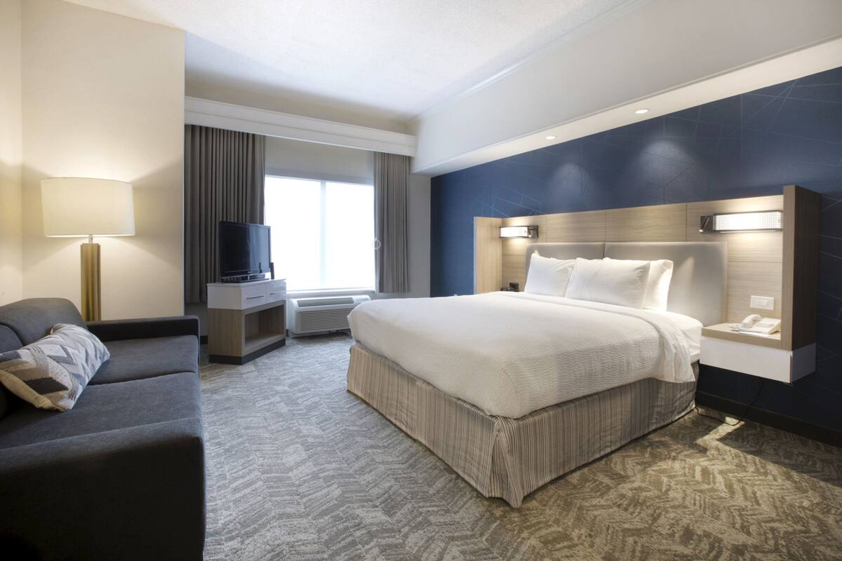 A hotel room with a large bed, a desk with a flat-screen TV, a sofa, and a large window with curtains.